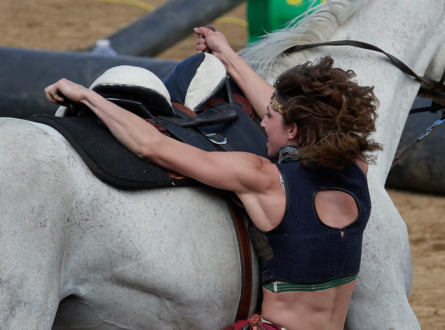 A woman with curly hair and wearing a dark sleeveless top performs trick riding, under the belly of a white horse at an outdoor riding area.