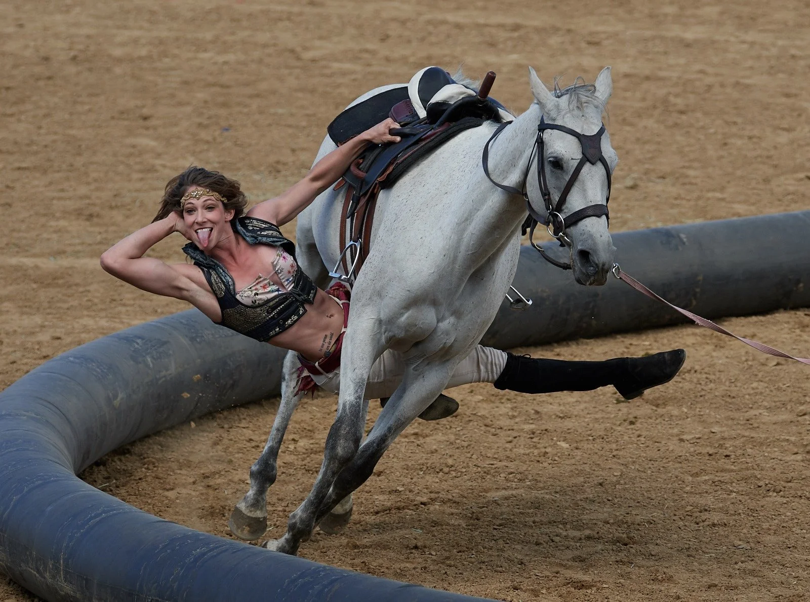 A woman in a colorful top and beige shorts is riding a white horse, passing under the horse performing trick riding. She is sticking out her tongue and smiling, with her left arm outstretched and her right hand holding the saddle.