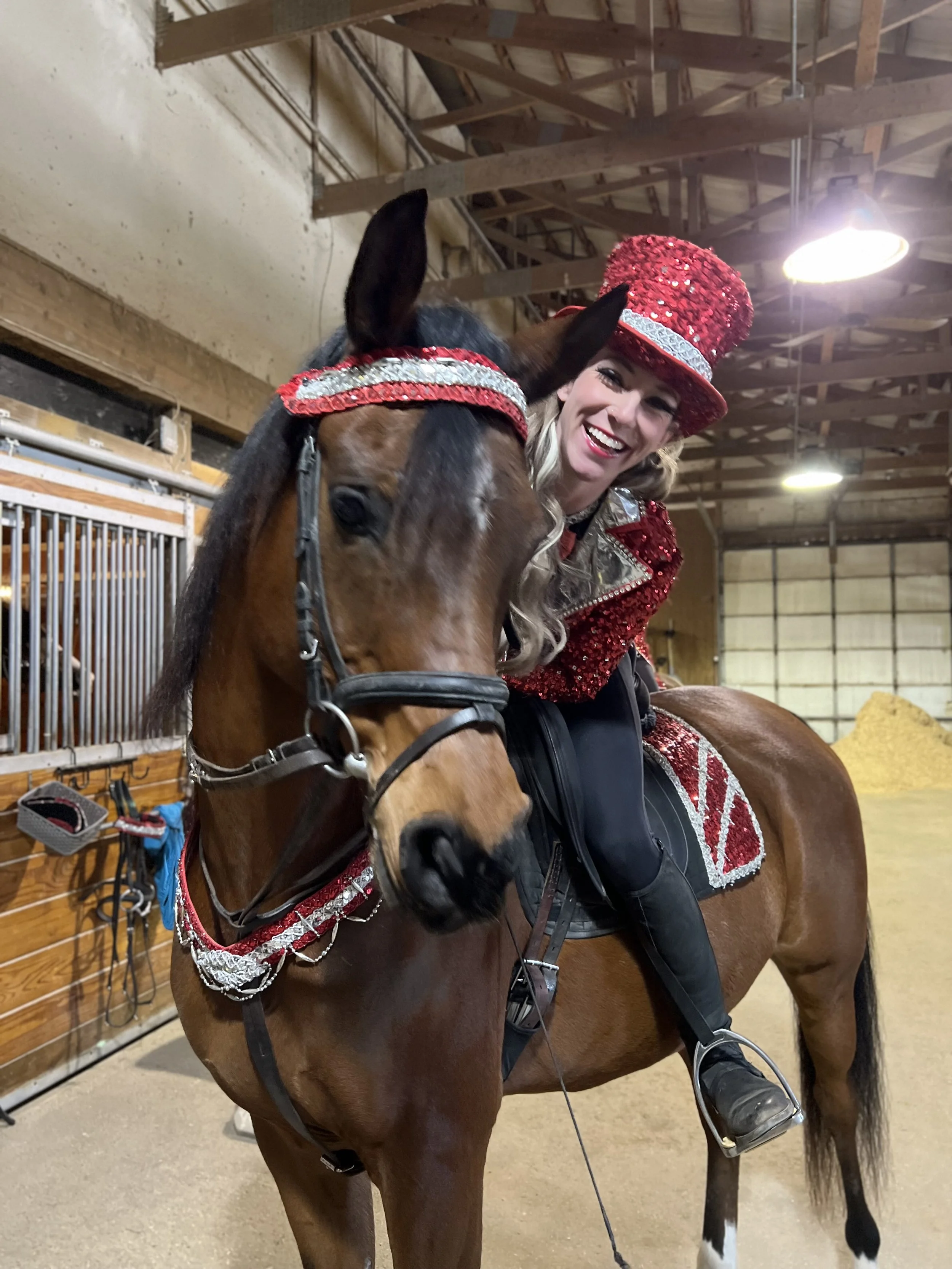 Woman dressed in a red and silver sparkly outfit with a matching top hat, riding a brown horse with a matching red and silver headband and saddle in a stable.