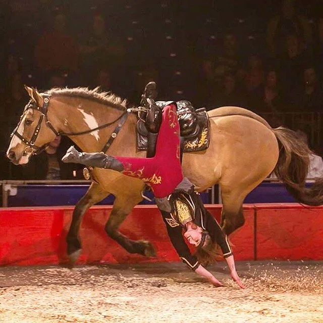 A woman performing a death drag on a horse in a circus arena with a red barrier and a dark background.