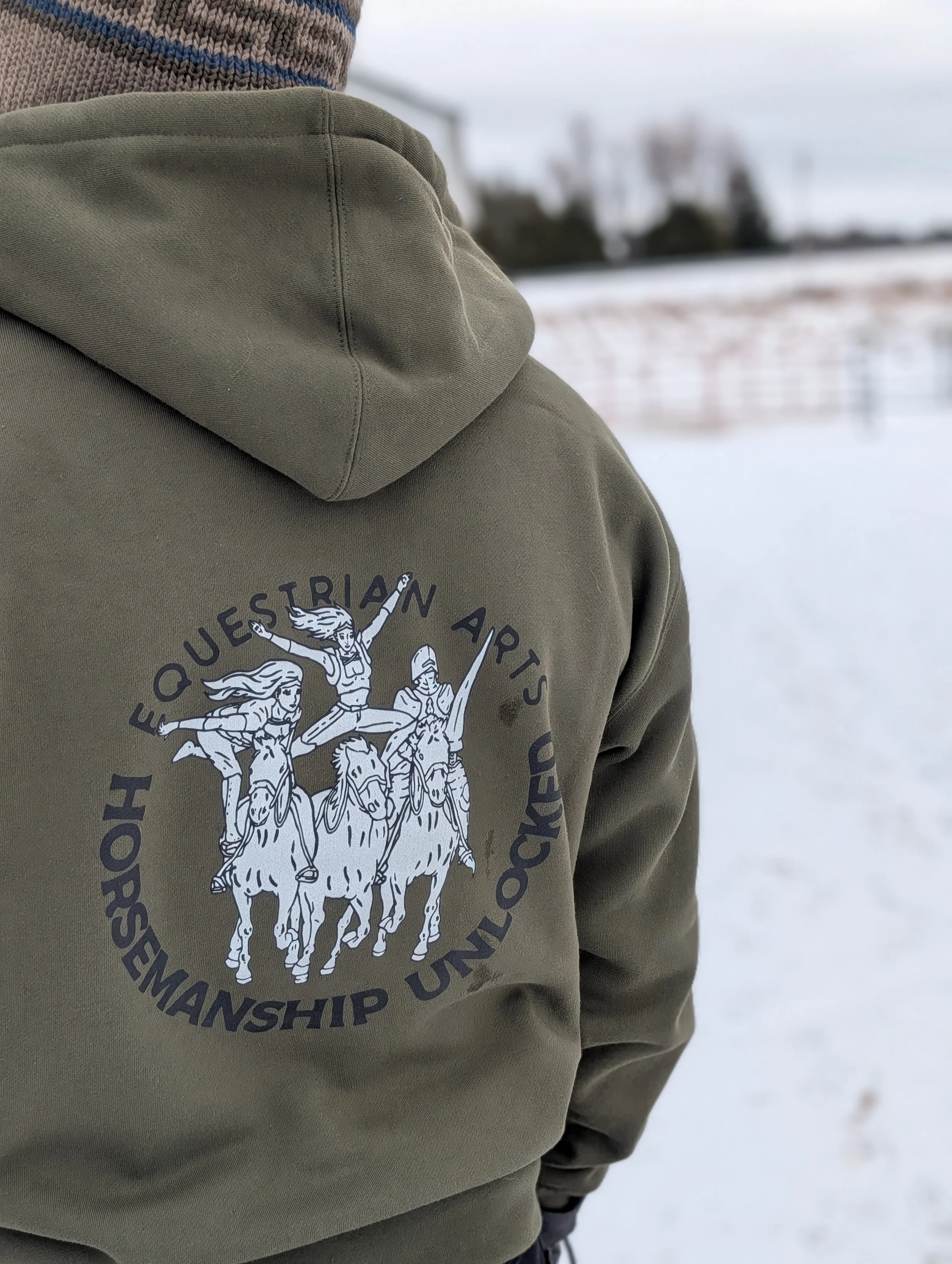 Close-up of a person wearing a green hoodie with a Questrian Arts Horsmanship Unlocked logo, featuring three women on horses, in an outdoor snowy landscape.