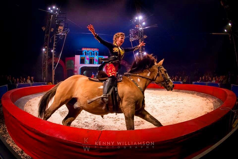 Circus performer riding a horse inside a ring, smiling and raising arms, with circus tent and audience in the background.