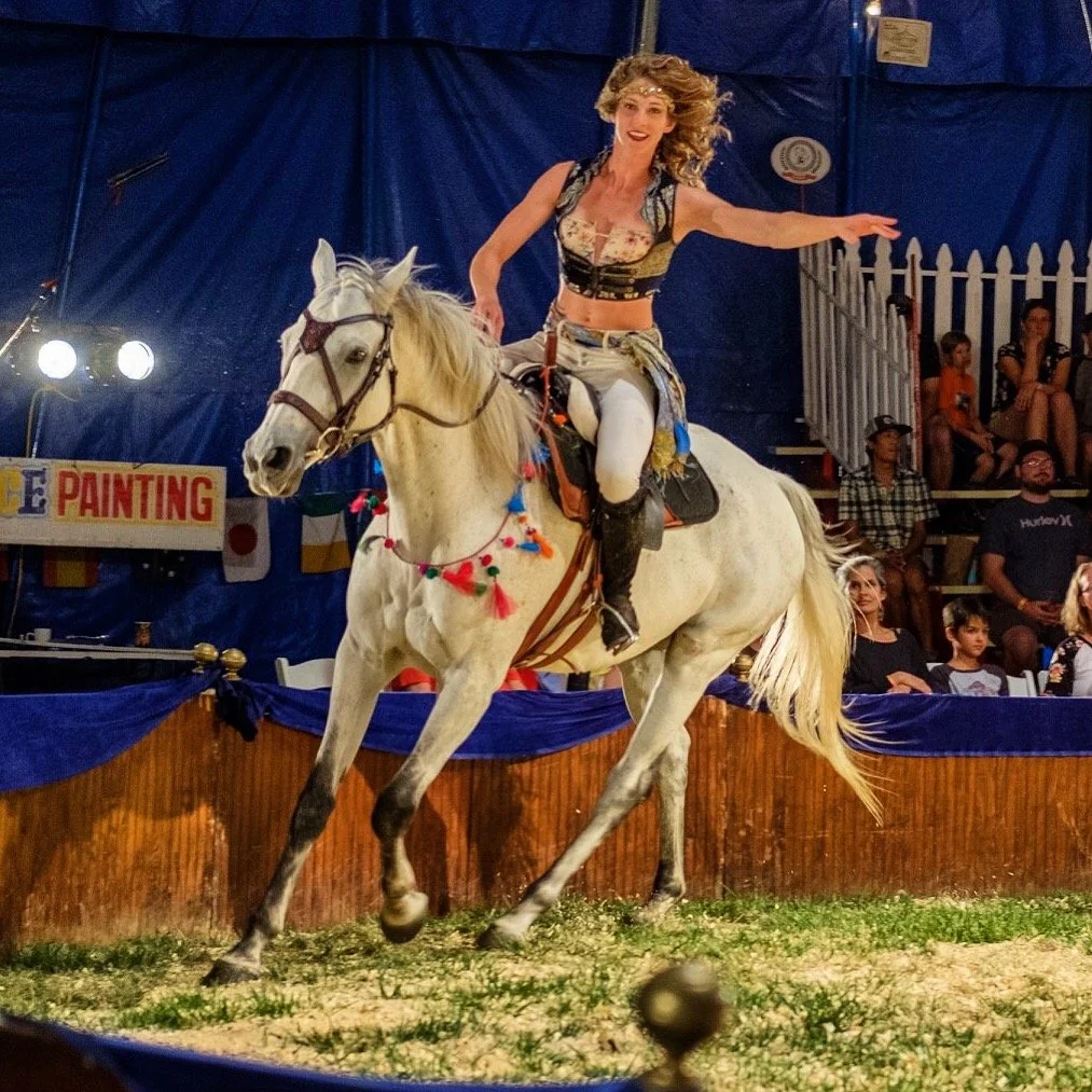 A woman riding a white horse inside a circus tent. The woman is wearing a top and white pants, and the horse has colorful decorations on its bridle. There are spectators seated in the background.