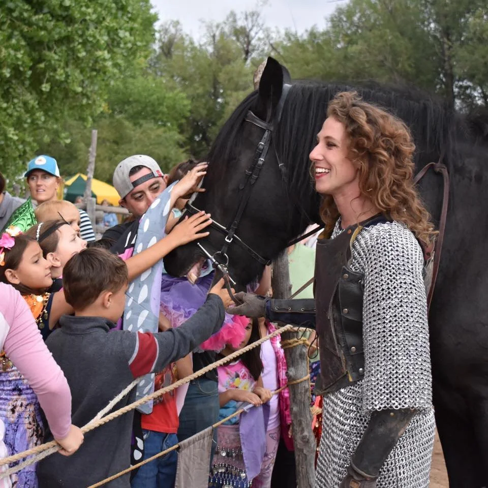 A woman with curly hair smiles at children reaching out to pet a black horse, with trees and a cloudy sky in the background.