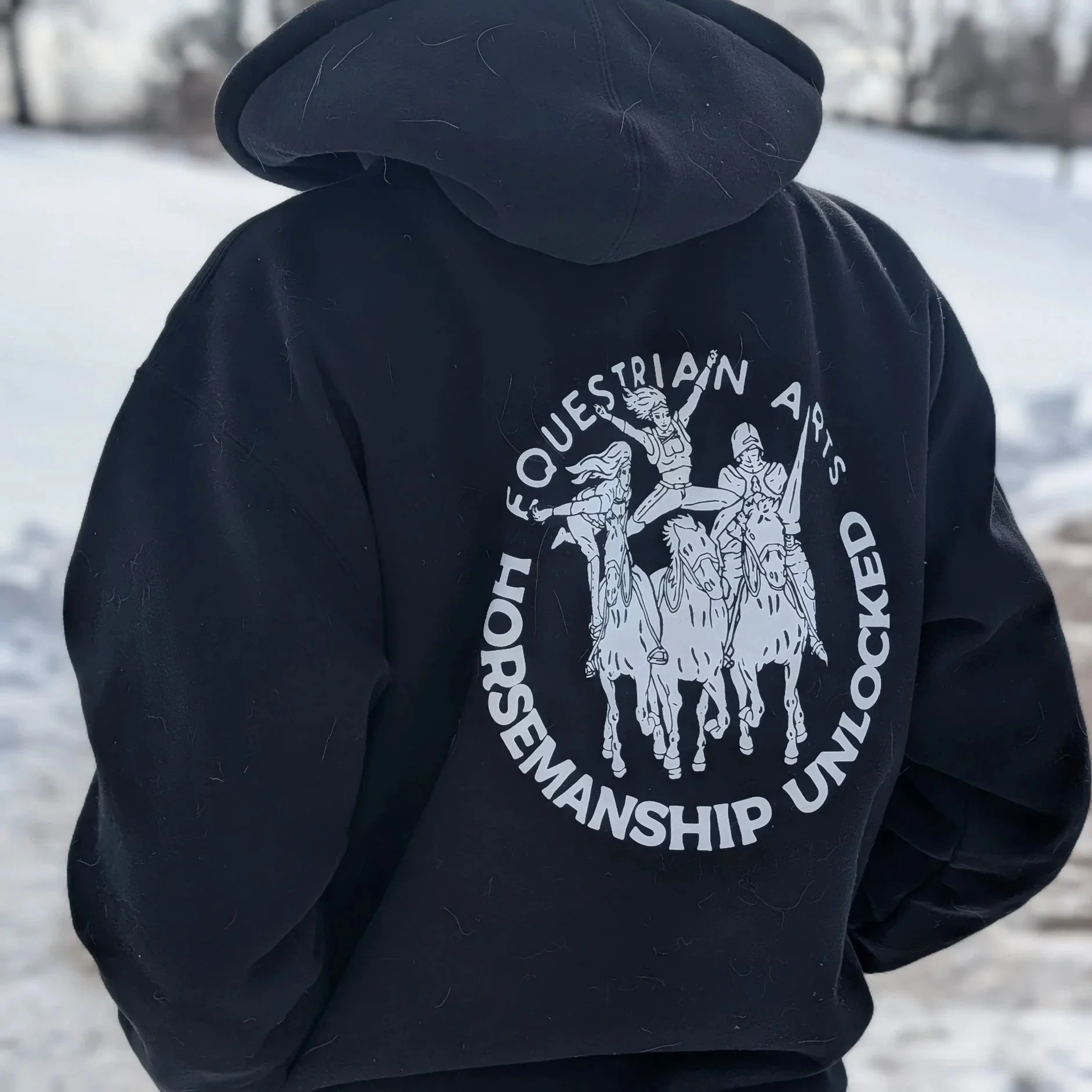 Person wearing a black hoodie with a design of three horse riders and the text 'Horsewoman Unrocked' and 'Foesquetrian Arts' on the back, standing outdoors in a snowy landscape.