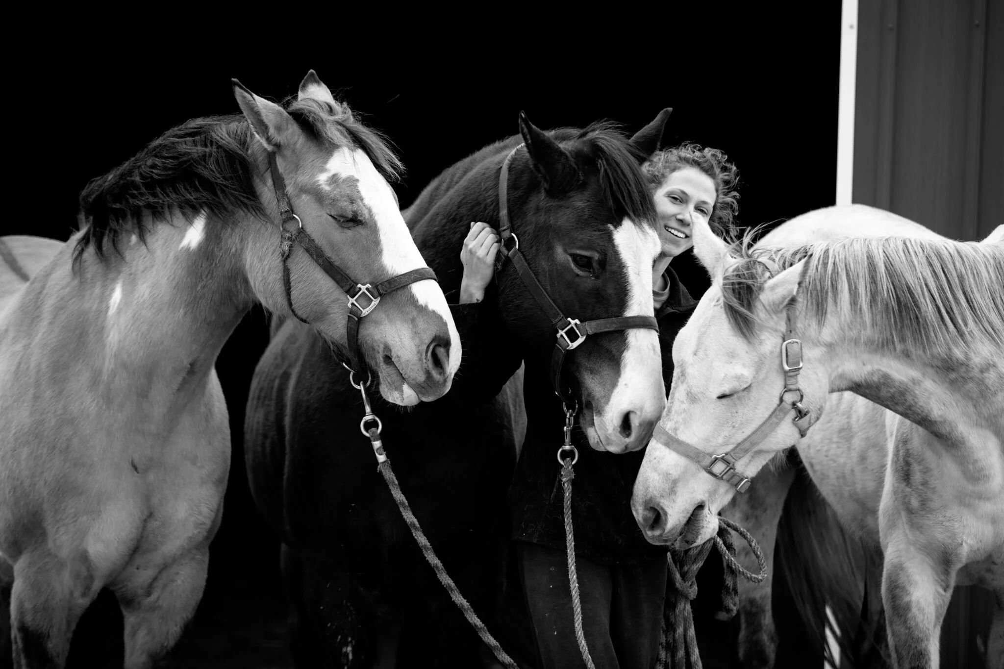 Woman smiling and hugging three horses in a stable, black and white photo.