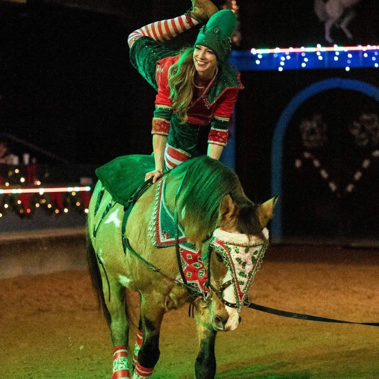 A woman in Christmas-themed clothing riding a decorated horse at night. She is smiling and wearing a green beanie, red and green jacket, and striped socks.