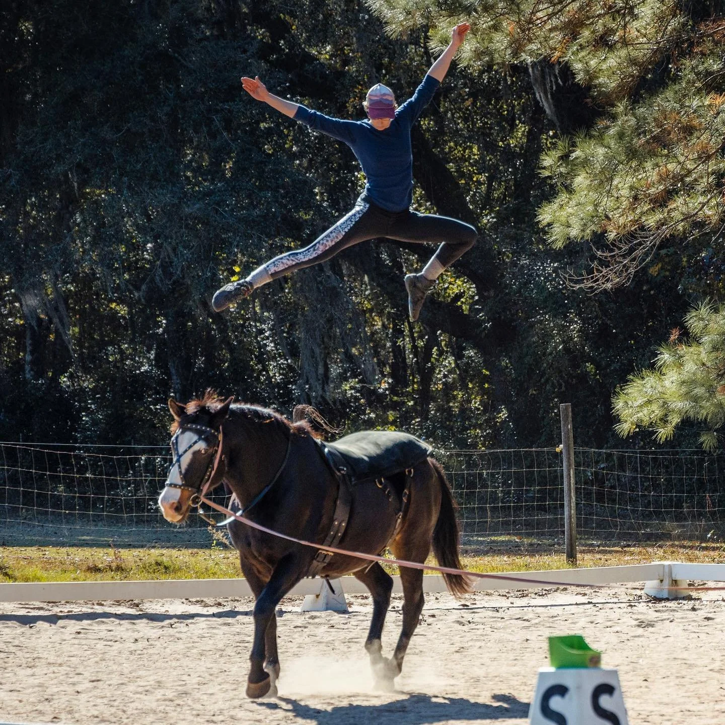 A person doing a jump in mid-air above a galloping vaulting horse on a sandy outdoor riding arena, with a backdrop of trees and a wire fence.