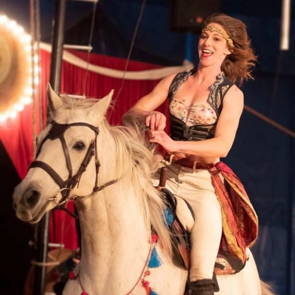 A woman with curly hair, wearing a headband and a colorful top, laughing while riding a white horse inside a circus tent with red curtains.