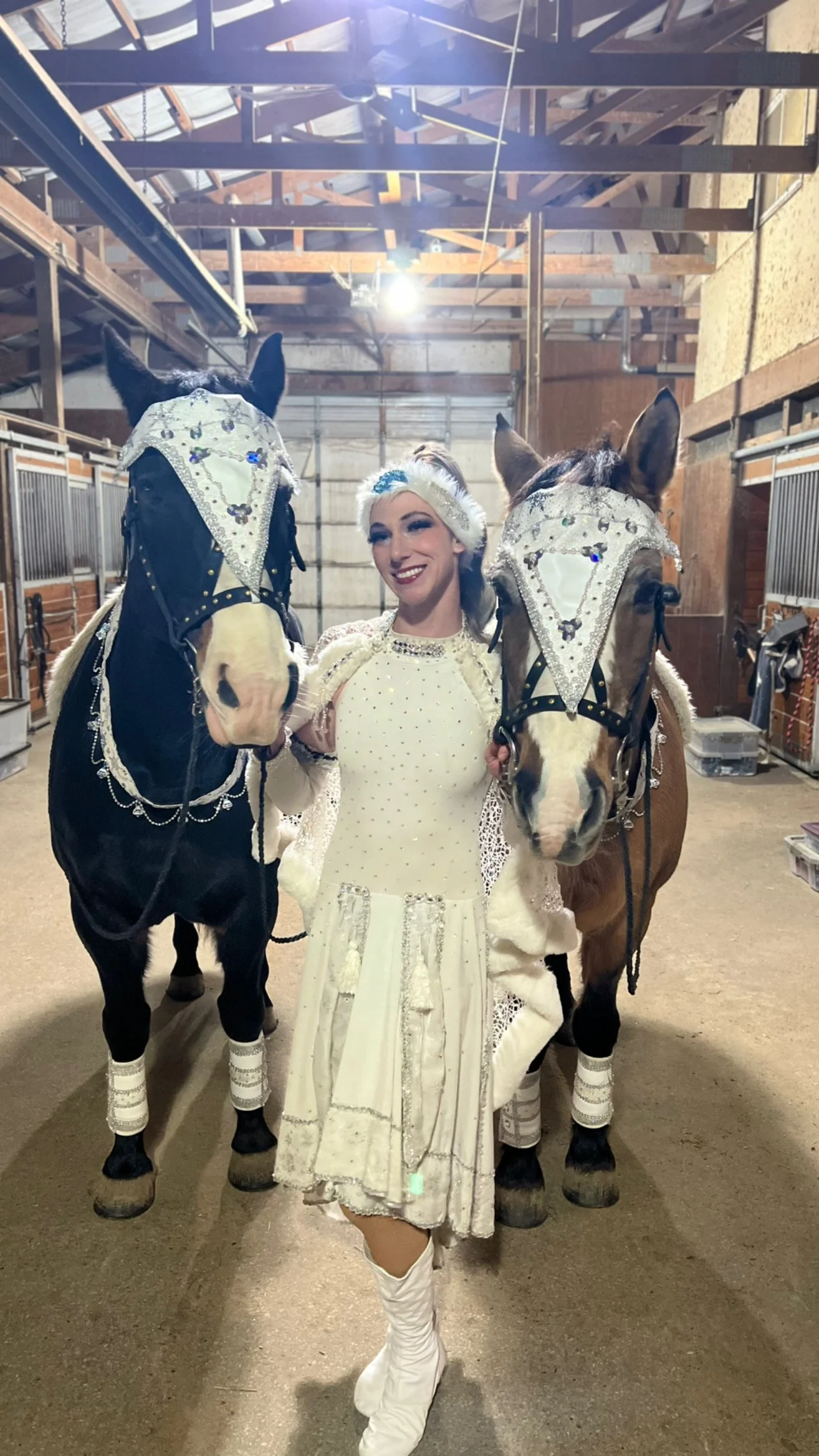 A woman dressed in a white costume with sequins, standing in a barn, holding the bridles of two decorated horses.
