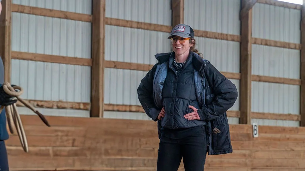 A woman standing indoors with wooden walls, wearing a black jacket, baseball cap, sunglasses, and smiling.