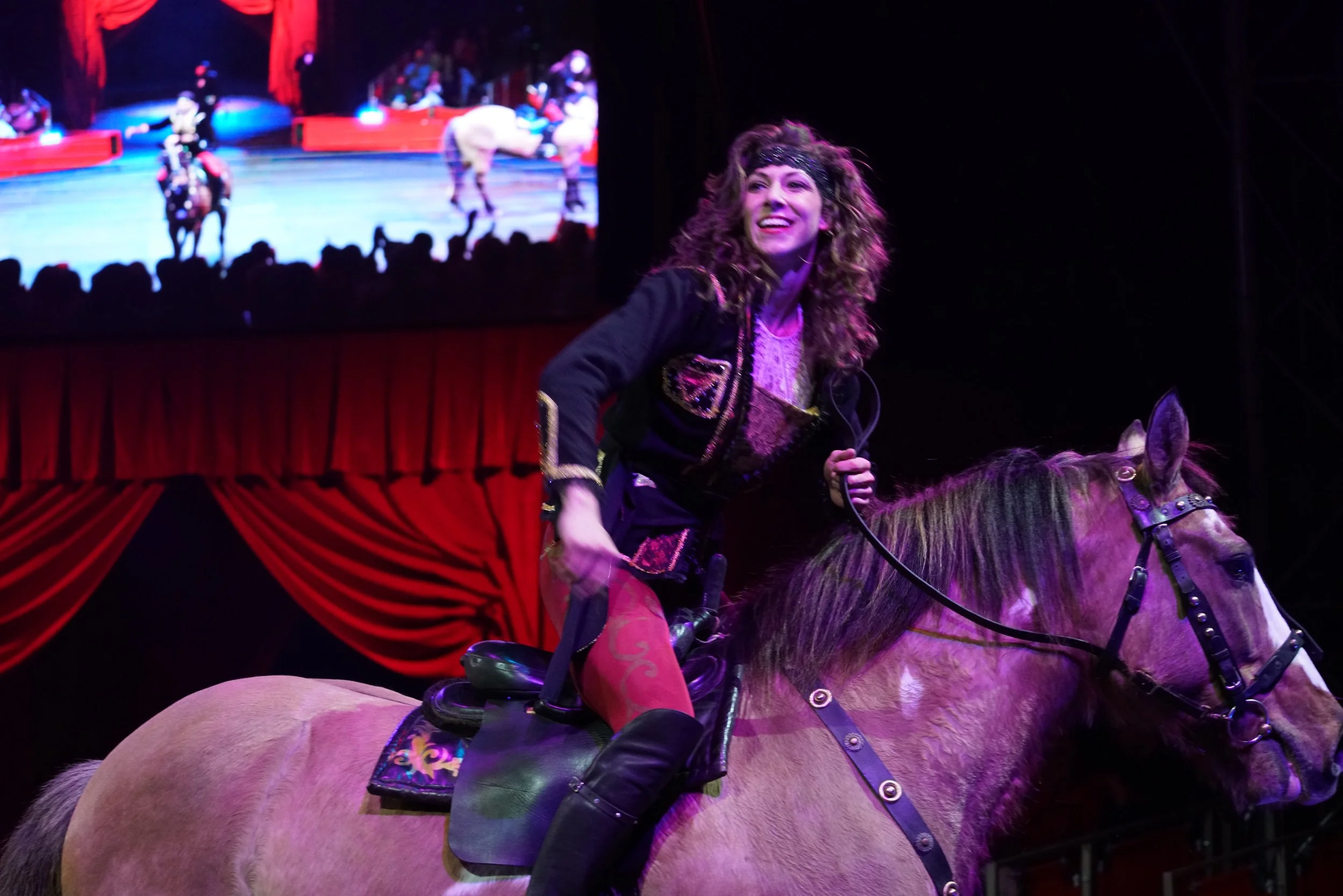 A woman with curly hair riding a horse in an indoor circus, smiling with audience in the background on a large screen.