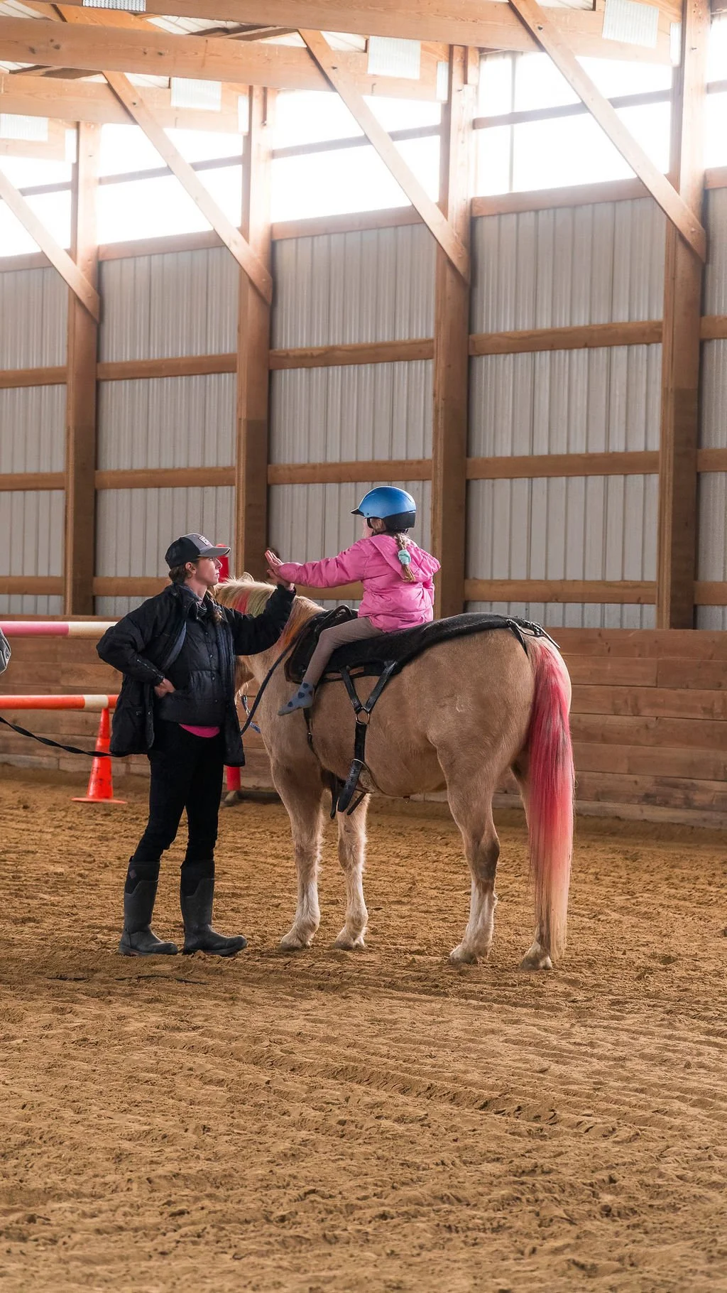 A young girl wearing a blue helmet sitting on a light brown horse in an indoor riding arena, while an adult woman in a black jacket and boots giving her a high-five.
