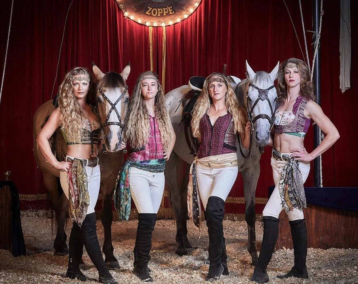 Four women dressed in circus or acrobatic costumes posing with two horses in a circus tent with red curtains and a sign that reads "Zoppë"