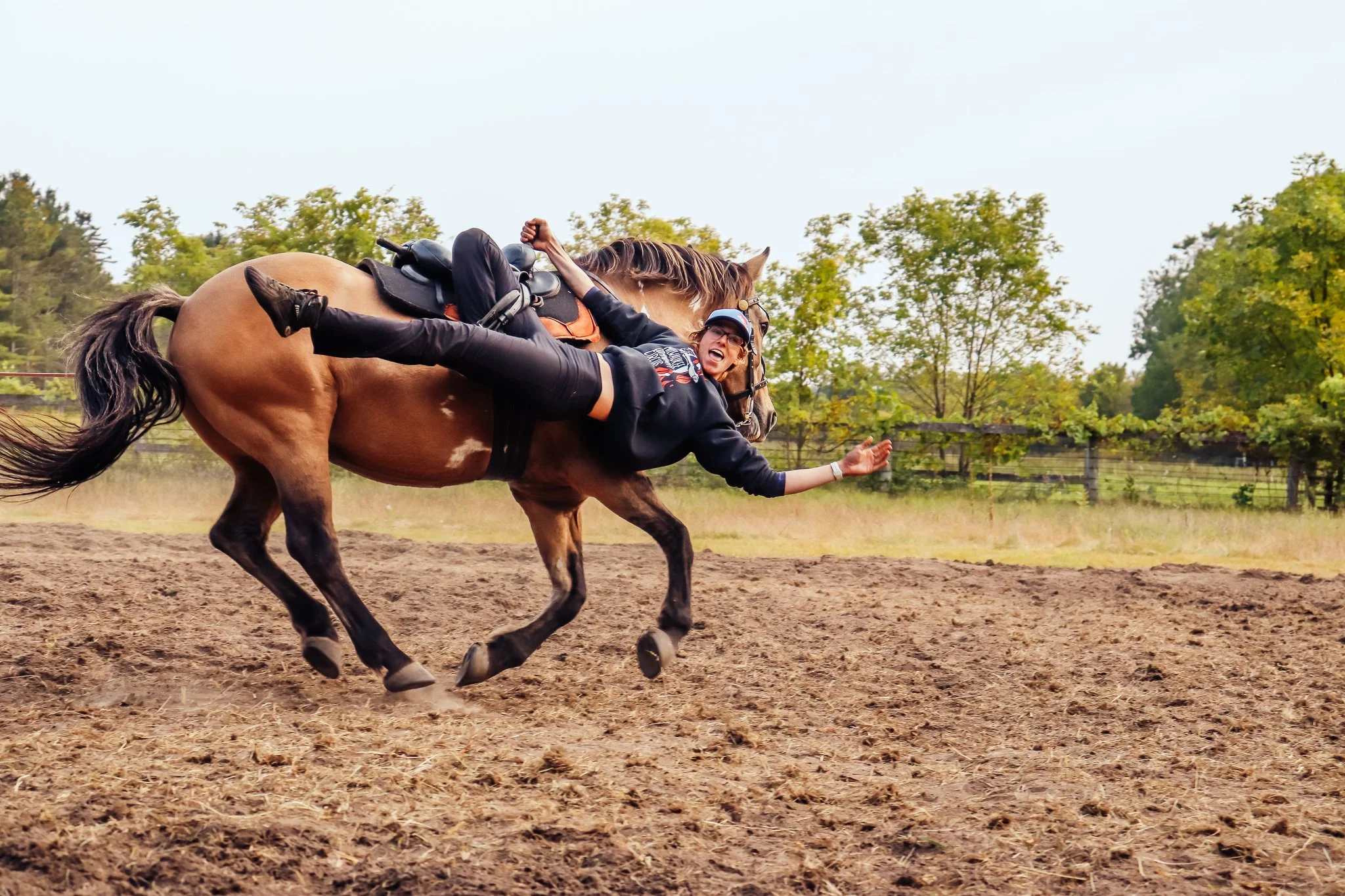 A woman trick riding a horse in an outdoor setting with trees and a fence in the background.