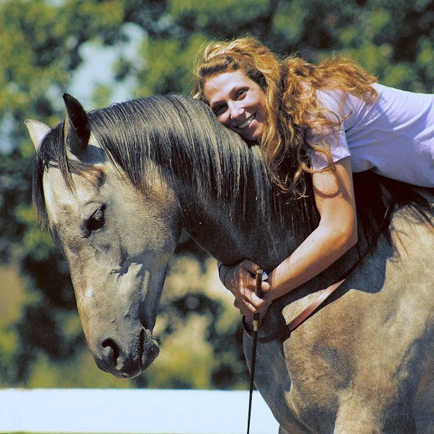 A young woman with curly red hair riding a gray horse outdoors during daytime, smiling and leaning forward.