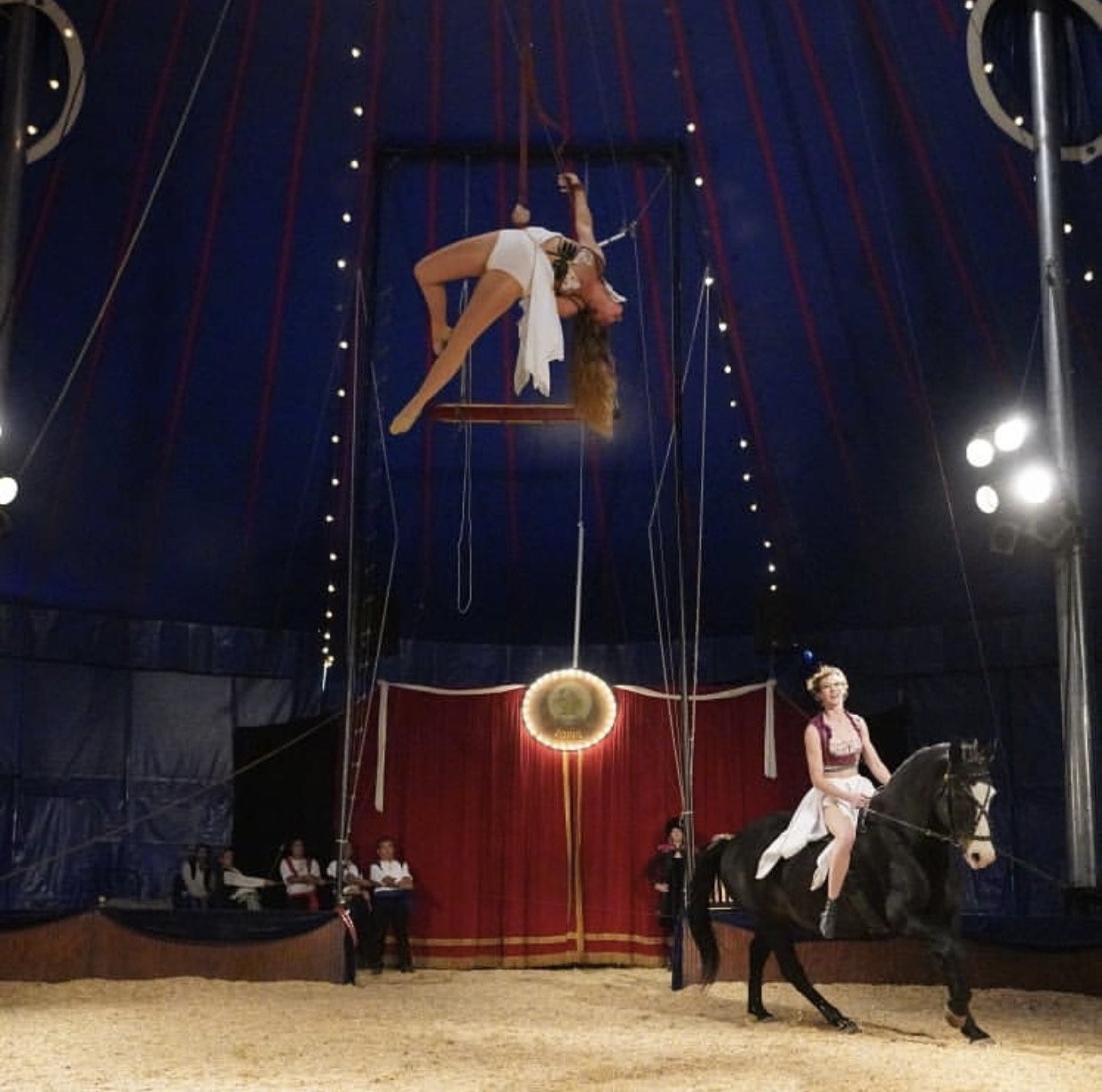 Circus performance with a woman hanging upside down from trapeze and a woman riding a horse on stage with a red curtain in the background.