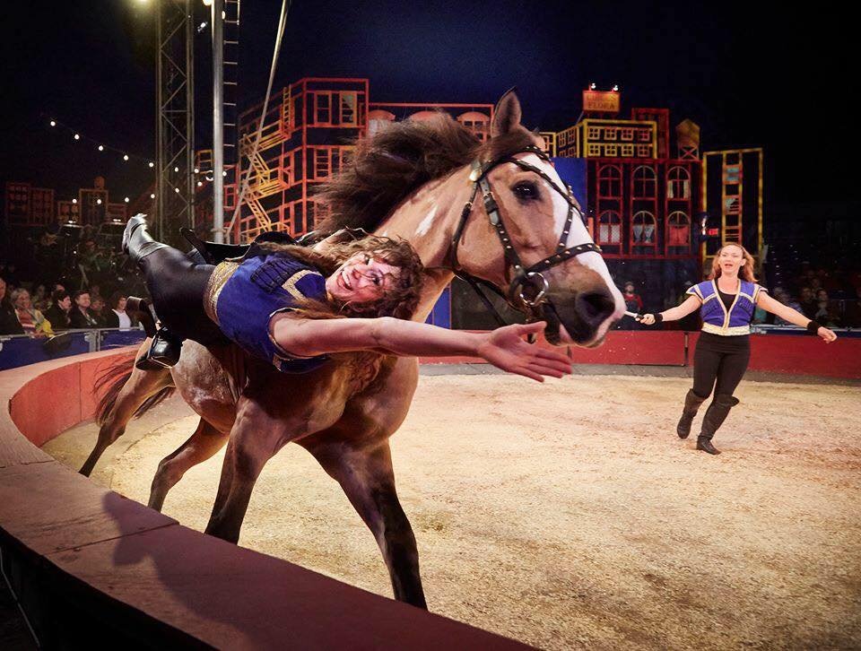 A woman riding a horse during a circus performance, with another performer in the background holding a stick in a circular arena.
