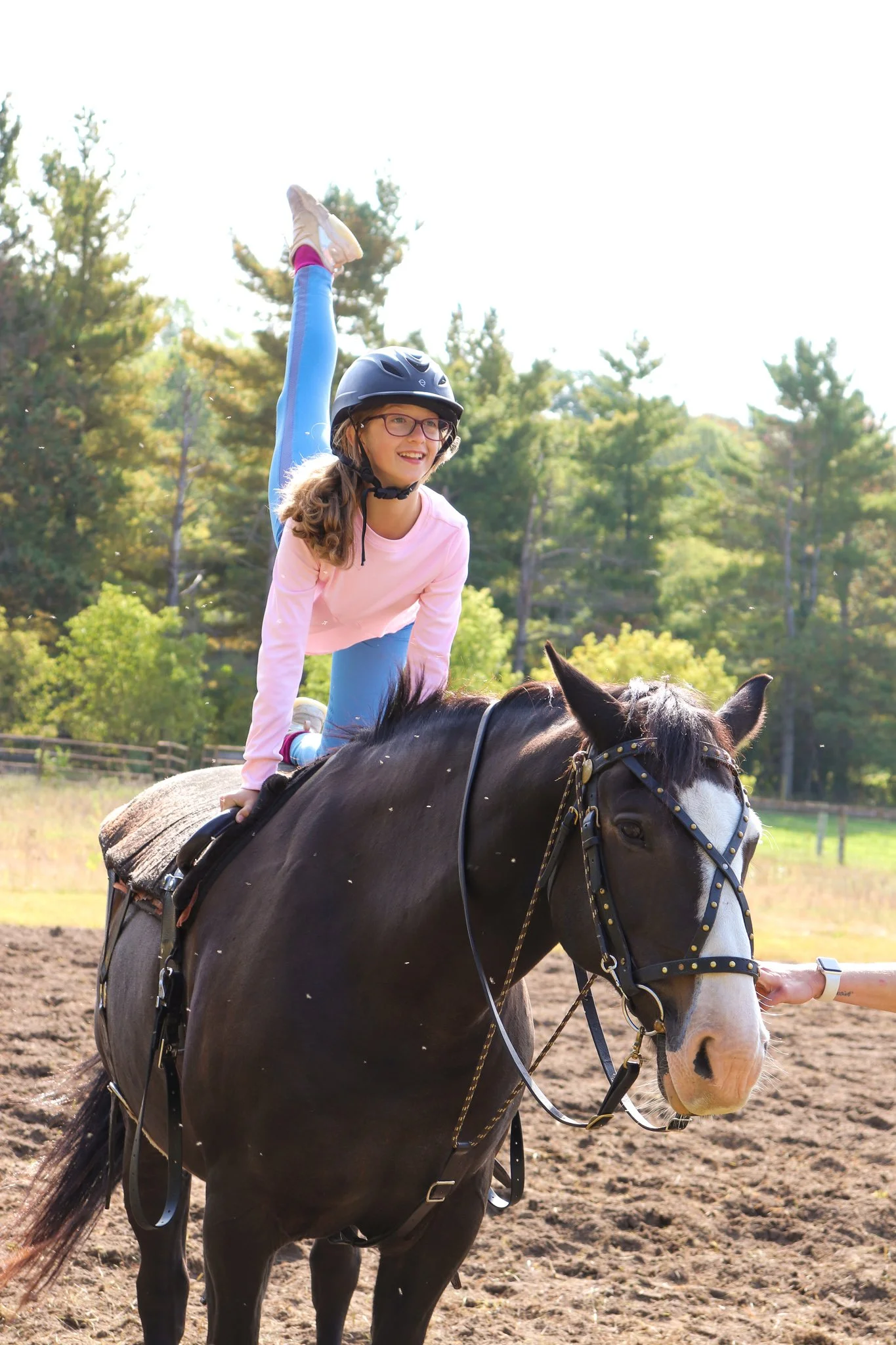 Young girl with glasses, pink sweatshirt, and blue leggings riding a black horse with white facial markings, wearing a helmet, outdoors on a sunny day.