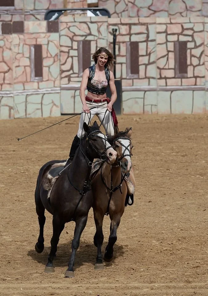 A woman is riding a horse indoors on a dirt floor, smiling and holding the reins, with a mural or painted wall in the background.
