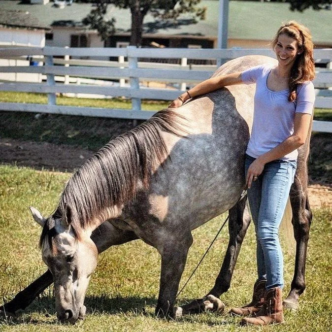 A woman with long red hair, wearing a white T-shirt, jeans, and brown boots, standing next to a gray horse with a darker mane, in a grassy area with a white fence and trees in the background.
