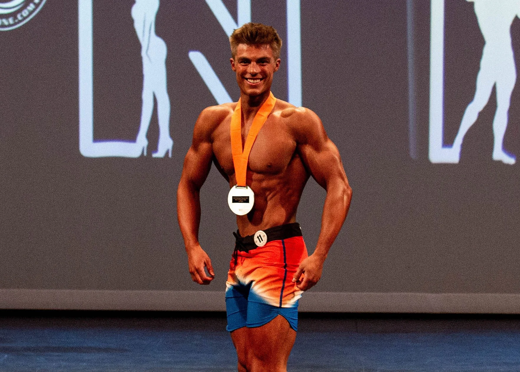 Men's Physique bodybuilding competitor on stage after earning a medal during a physique competition.