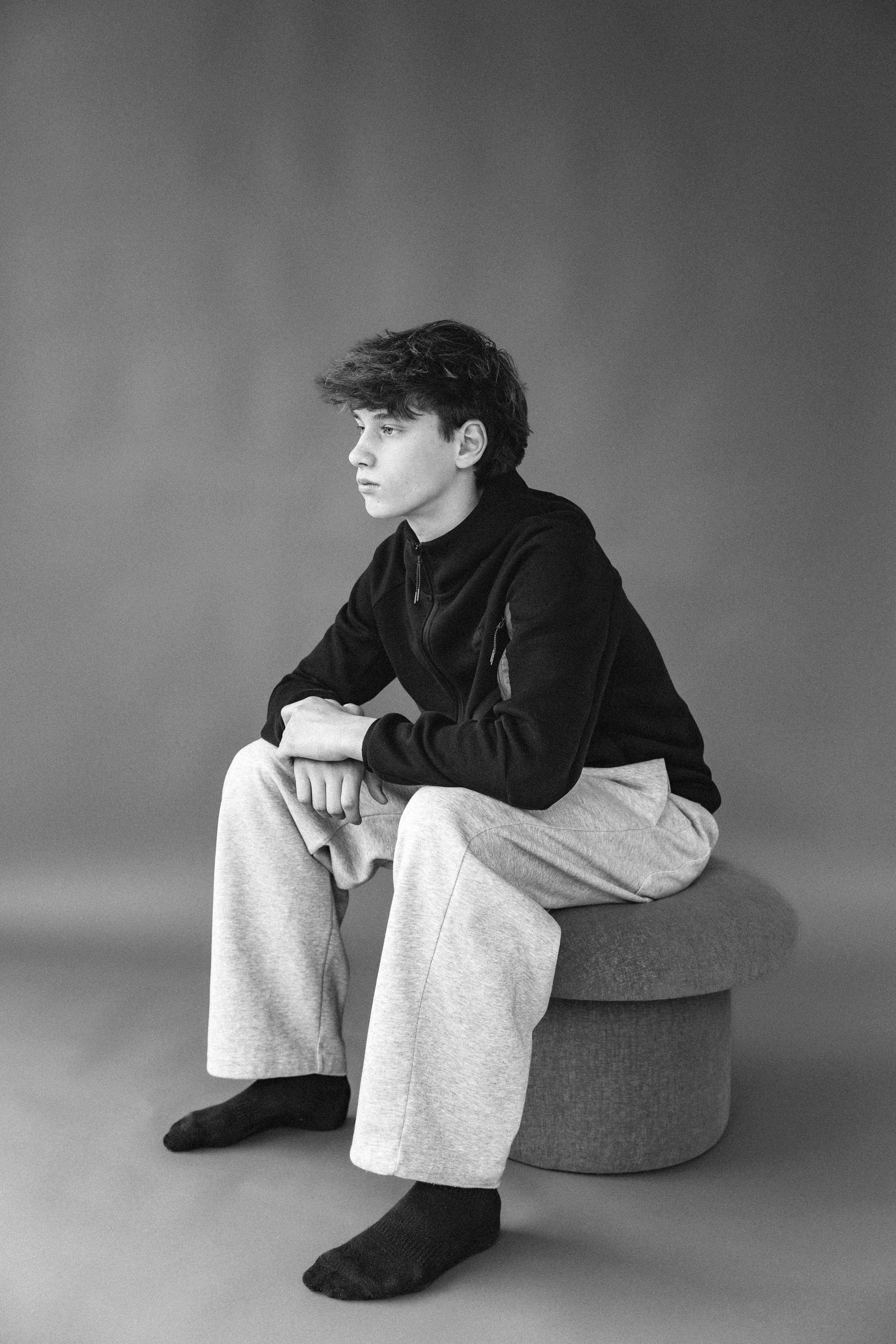 Black and white profile portrait of seated teen boy against seamless backdrop.