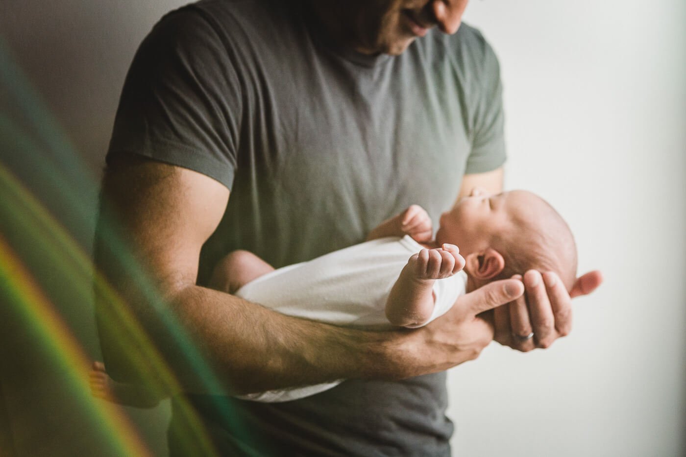 Crop of dad's strong arms holding newborn son while rainbow light leaks in.