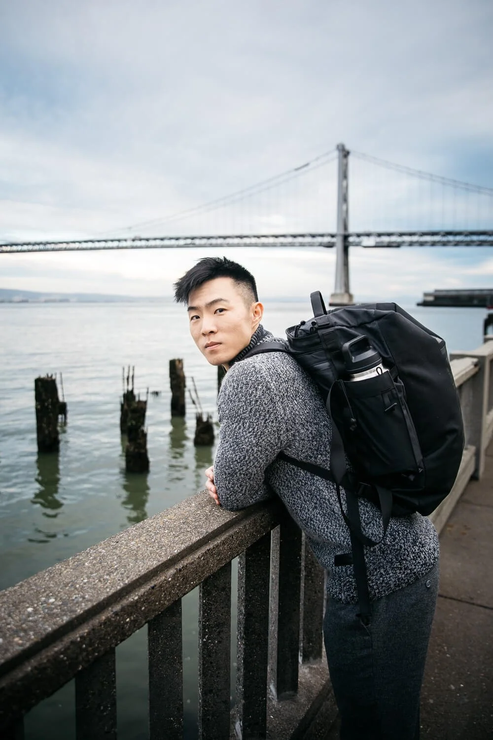 Asian man with large black backpack leans over railing with Bay Bridge in the background.