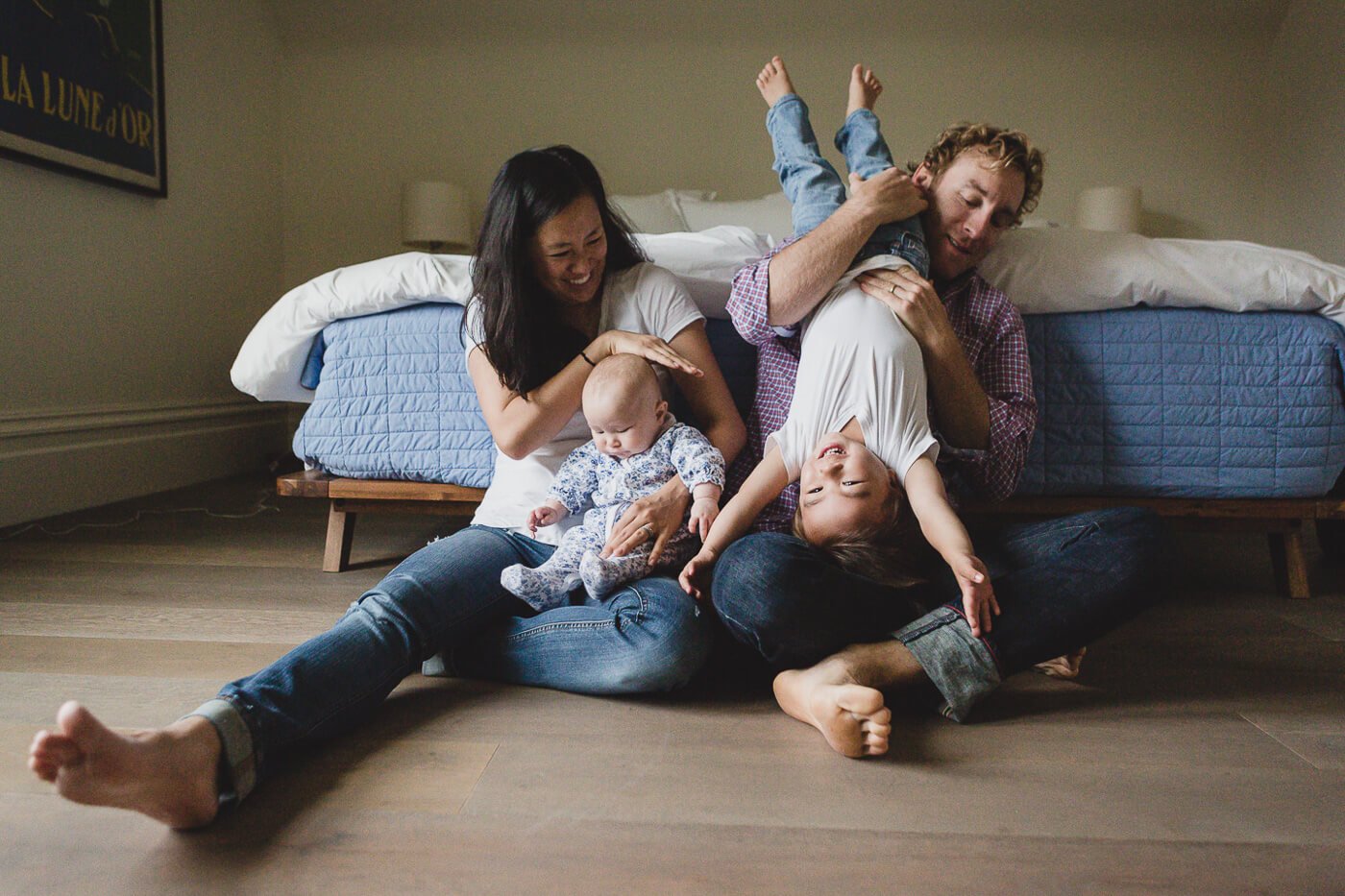 Family of four sits on bedroom floor while holding newborn and playing with toddler, taken at home during newborn lifestyle session.