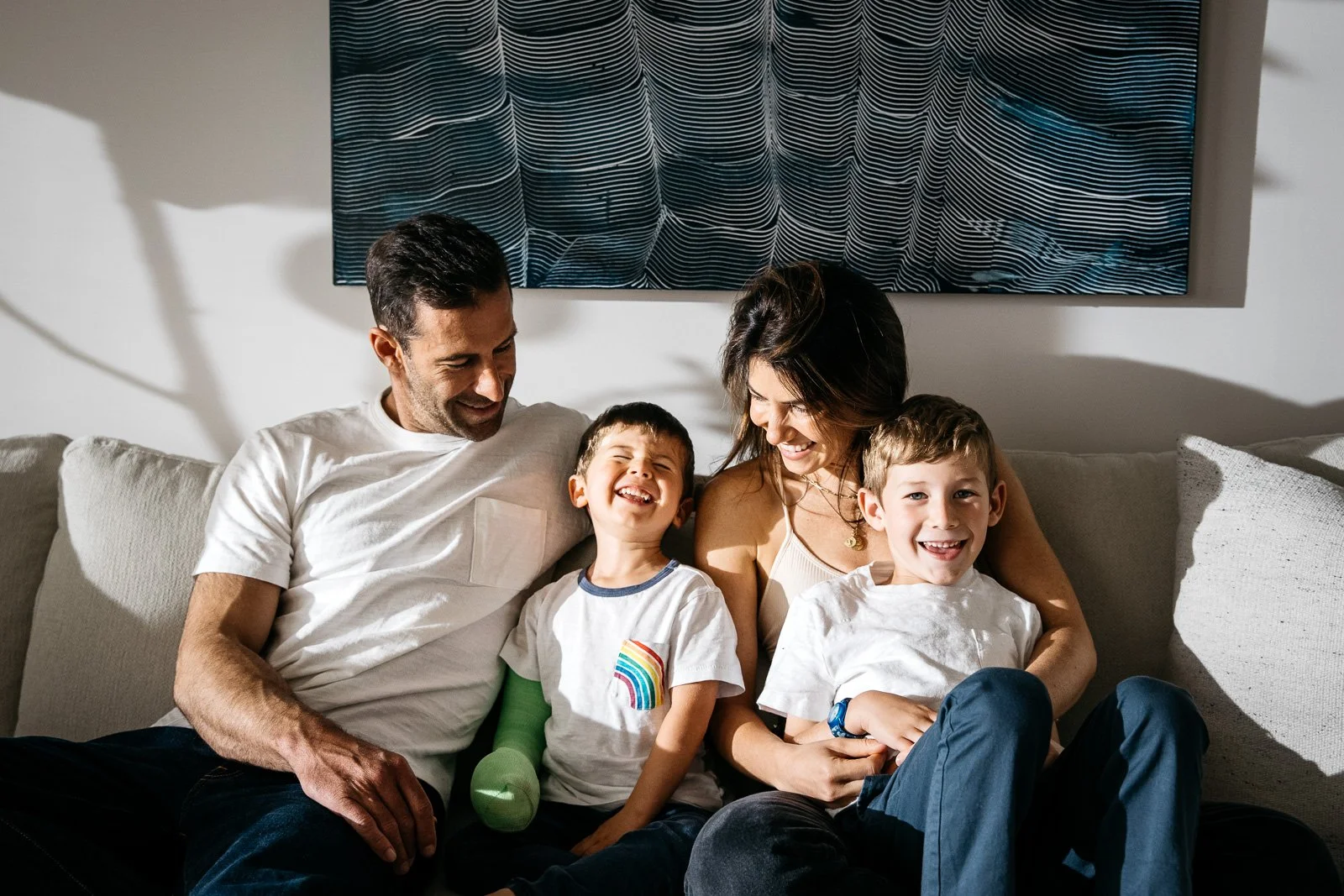 Family with two little boys sits on white counch with navy blue paiting behind them, laughing as sunset light streams in from the left.