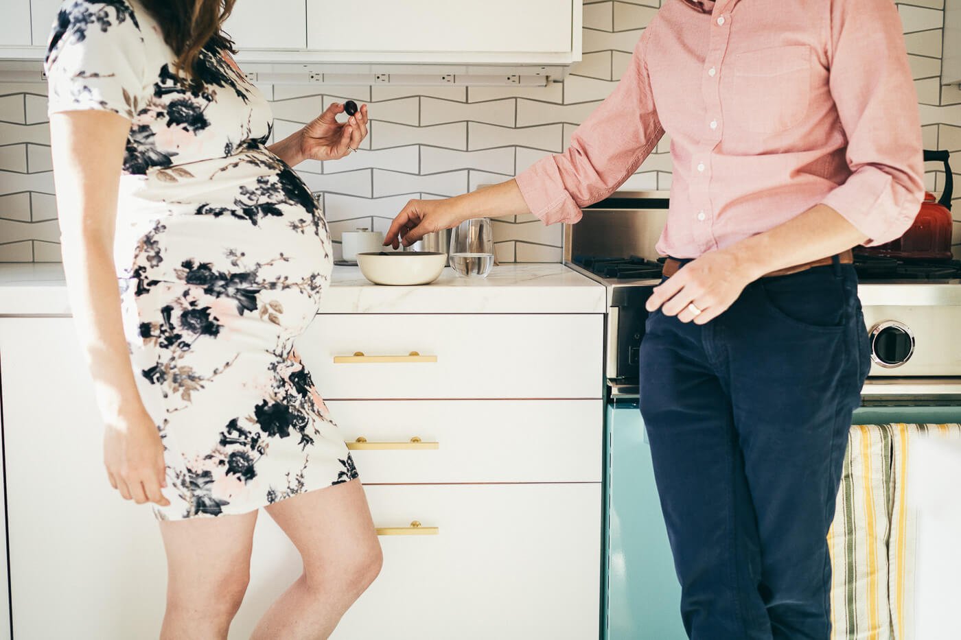 Profile midsection crop of pregnant belly across from partner as they stand in kitchen and eat cherries.