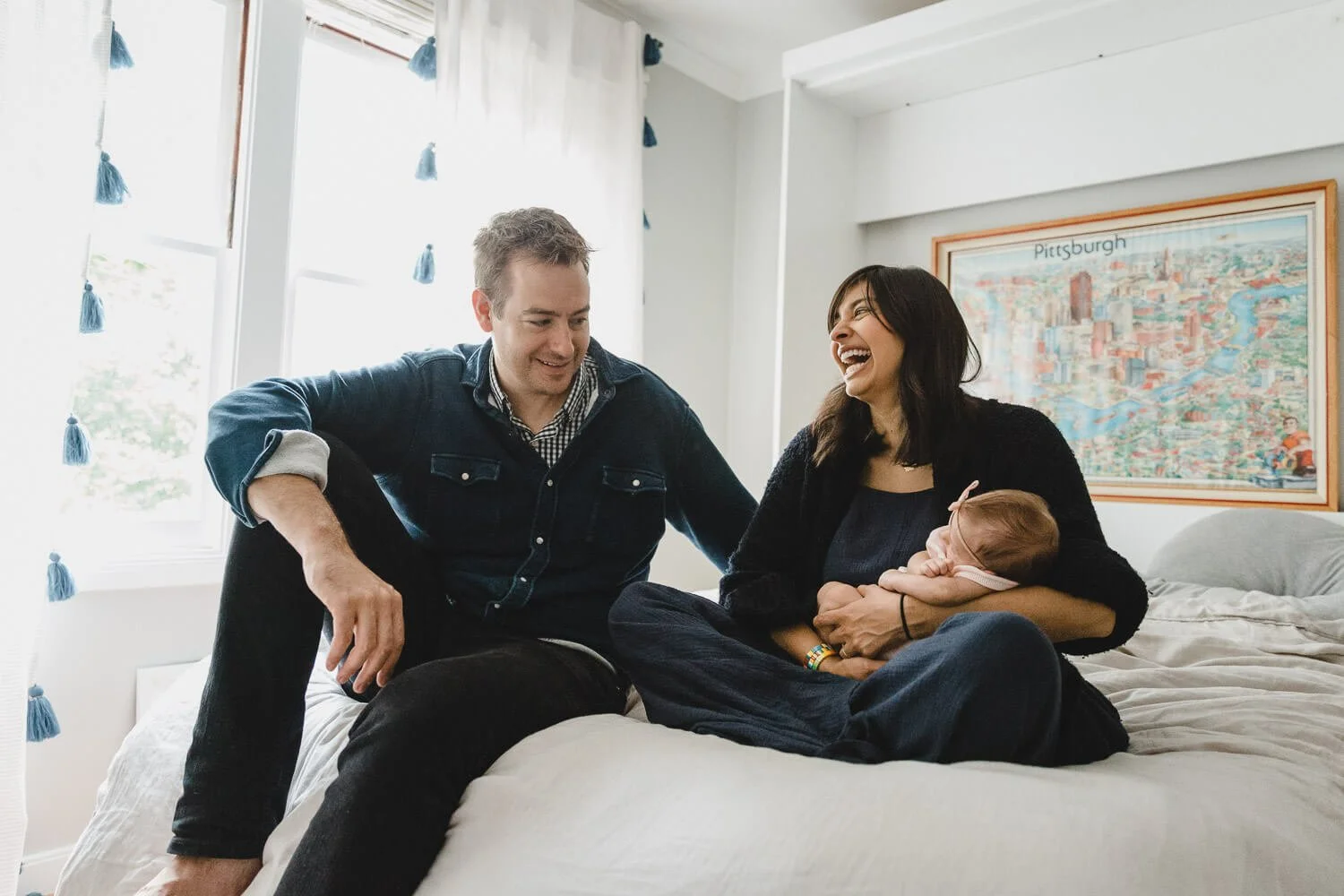 Mom and dad sit on white bed, laughing, while holding sleeping newborn baby, with sunlight streaming in the large window next to them. Taken during newborn lifestyle session at home in Berkeley.