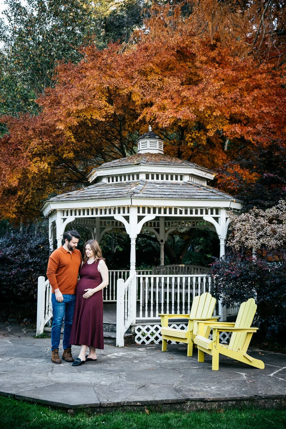 Pregnant mama and husband stand facing each other in front of white gazebo with giant trees with fall color surround them. Shot during maternity session at Marin Art and Garden Center.
