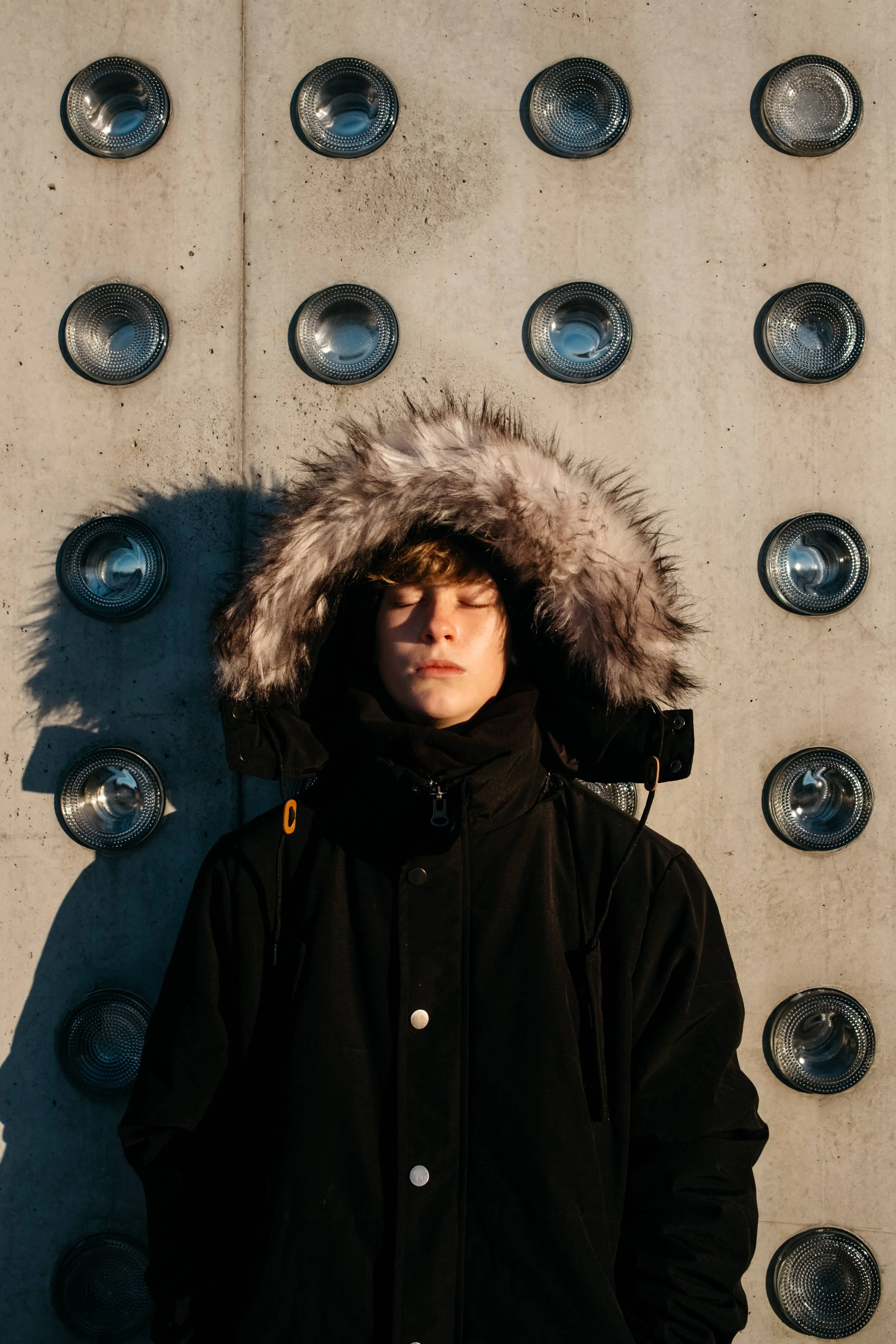 Boy in black coat and fur-lined hood stands against cement wall in the sunshine.