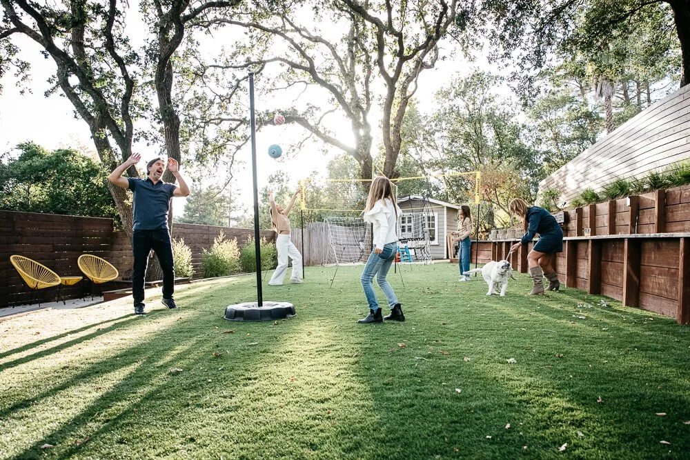 Dad and daugther play tether ball in backyard, next to mom who is playing with white lab puppy, while two sister behind them play volleyball.