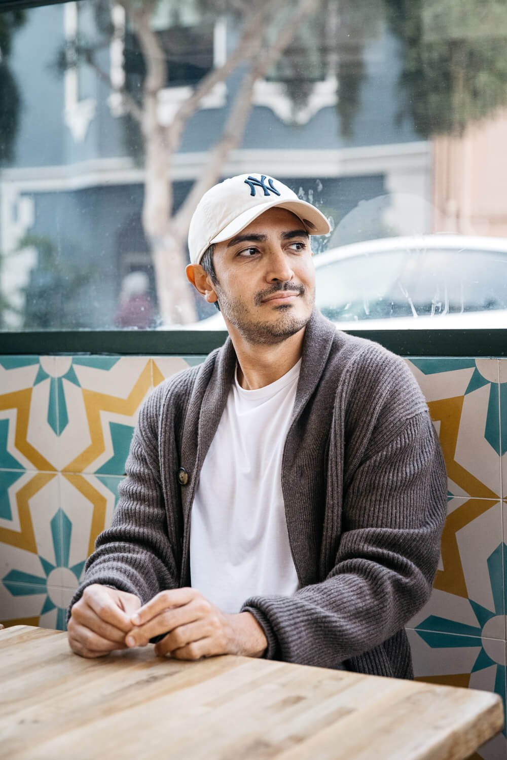 Man wearing white Yankees cap sits at cafe table with colorful tile seat back.
