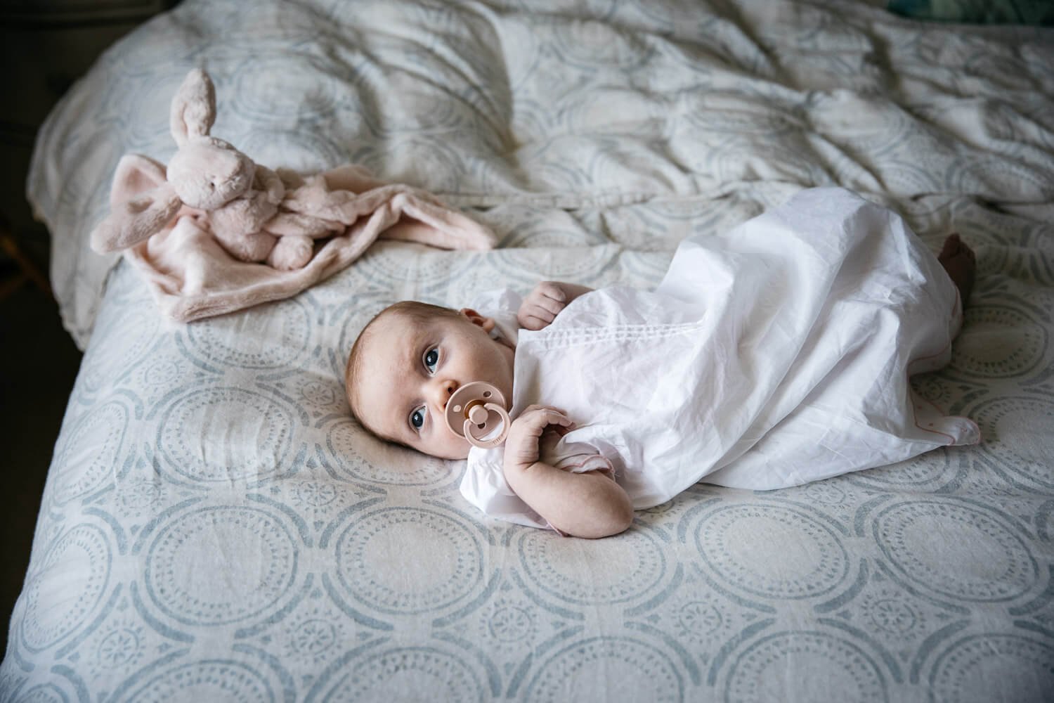 Newborn in white christening dress lies on bed with pink pacifier and pink stuffed bunny, taken at home in Orinda.