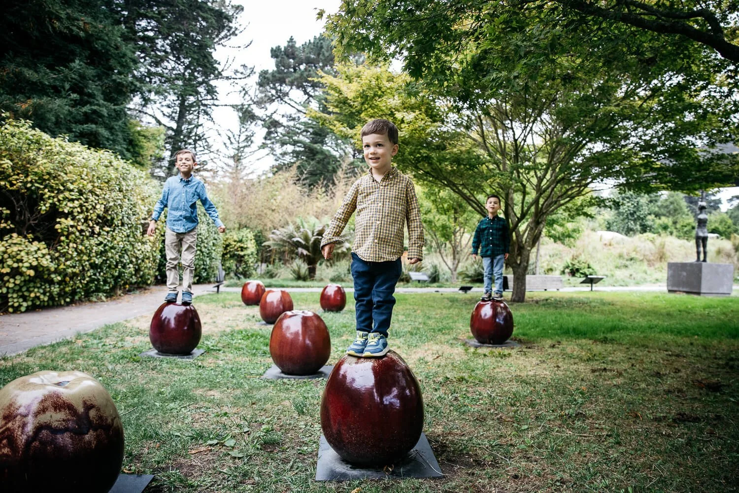 Three boys balancing on large decorative apple sculptures in the garden outside the de Young Museum during a San Francisco family mini session in Golden Gate Park.