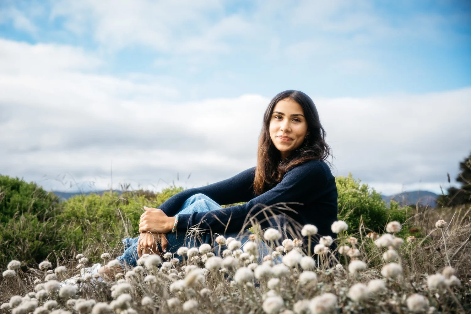 South Asian high school senior teen girl with navy sweater and jeans sits in field of white flowers at Lands End. She smiles at the camera with the wind in her hair. Taken during outdoor senior photo shoot.