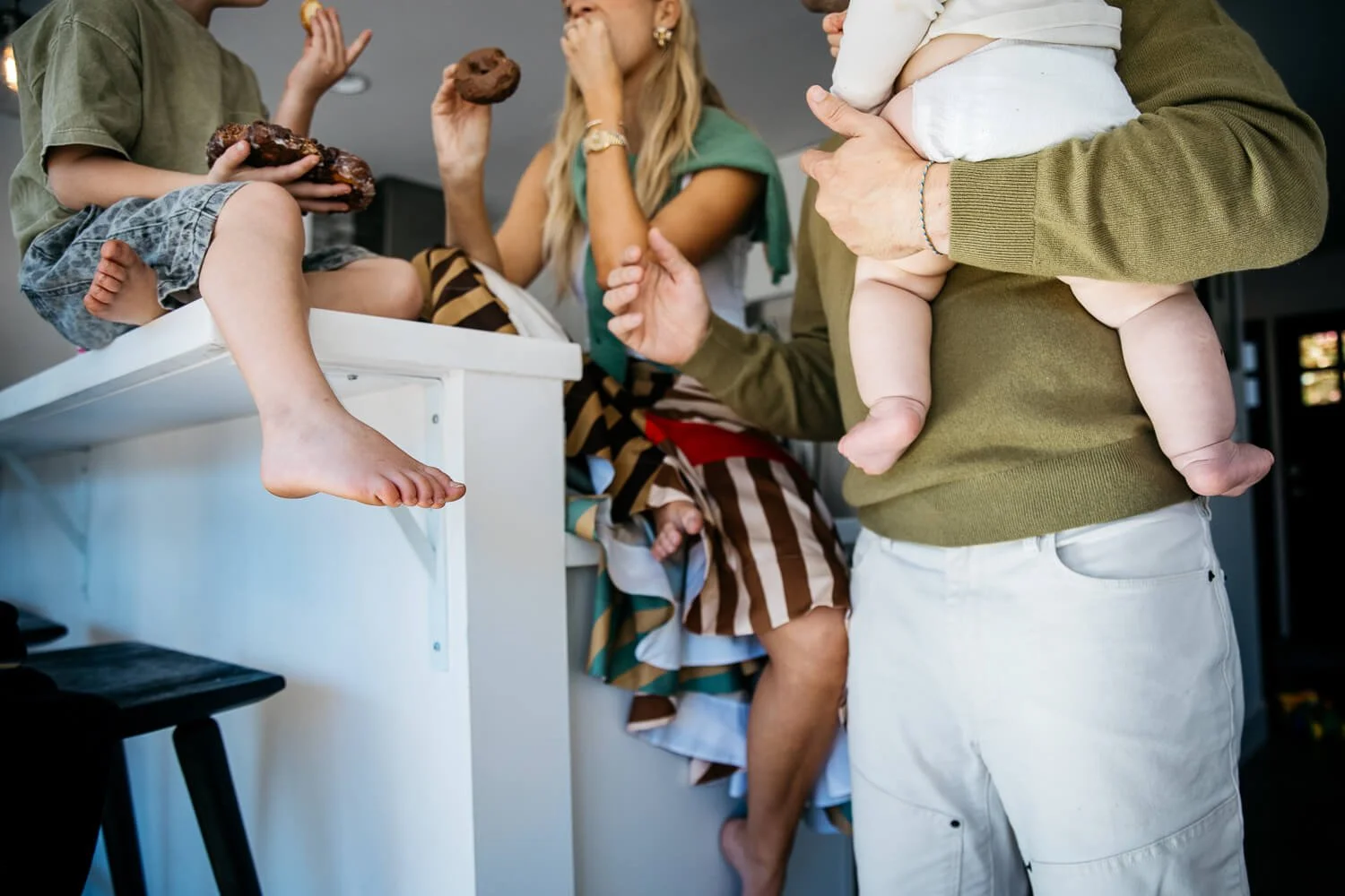 Family sits on kitchen  counter, eating donuts during the family photos session at home.