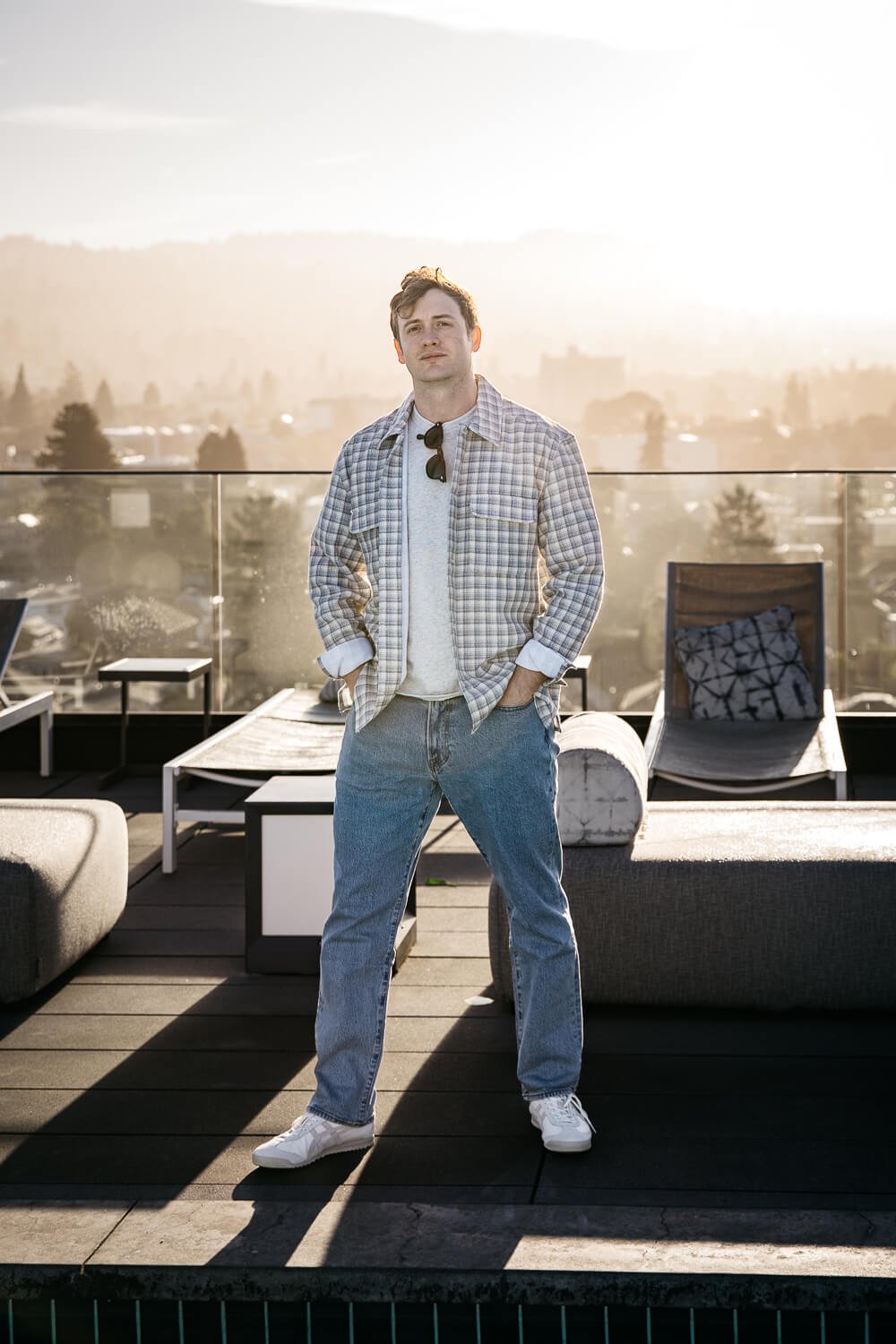 Man with white tee and checkered overshirt and jeans, stands on rooftop deck with the sunrise and Oakland hills behind him.