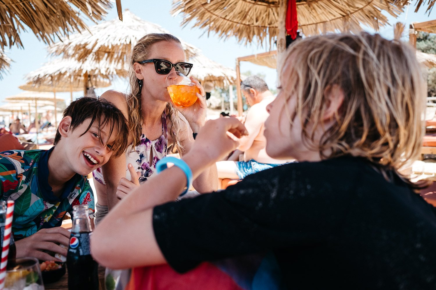 Mom drinks cocktail under beach unmbrella as two kids in bathing suits goof off next to her.