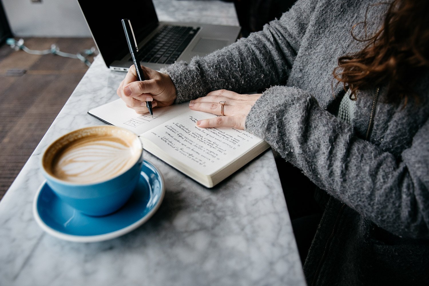  overhead shot of woman hand writing in journal next to blue coffe cup filled with cappuccino, shot at Home Coffee Raosters in San Francisco by brand photographer Allison Busch Photography 