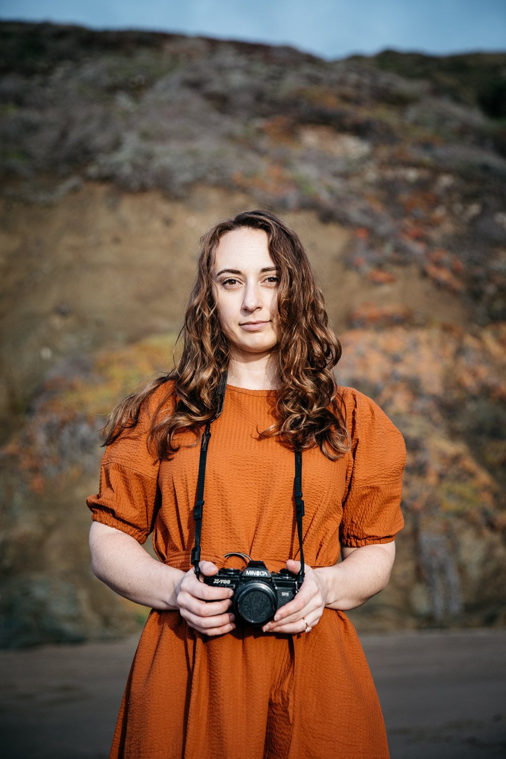  brand portrait of photographer Toni Toscano holding her Olympus camera with a cliff and sky behind her, shot at Baker Beach in San Francisco by brand photographer Allison Busch Photography 