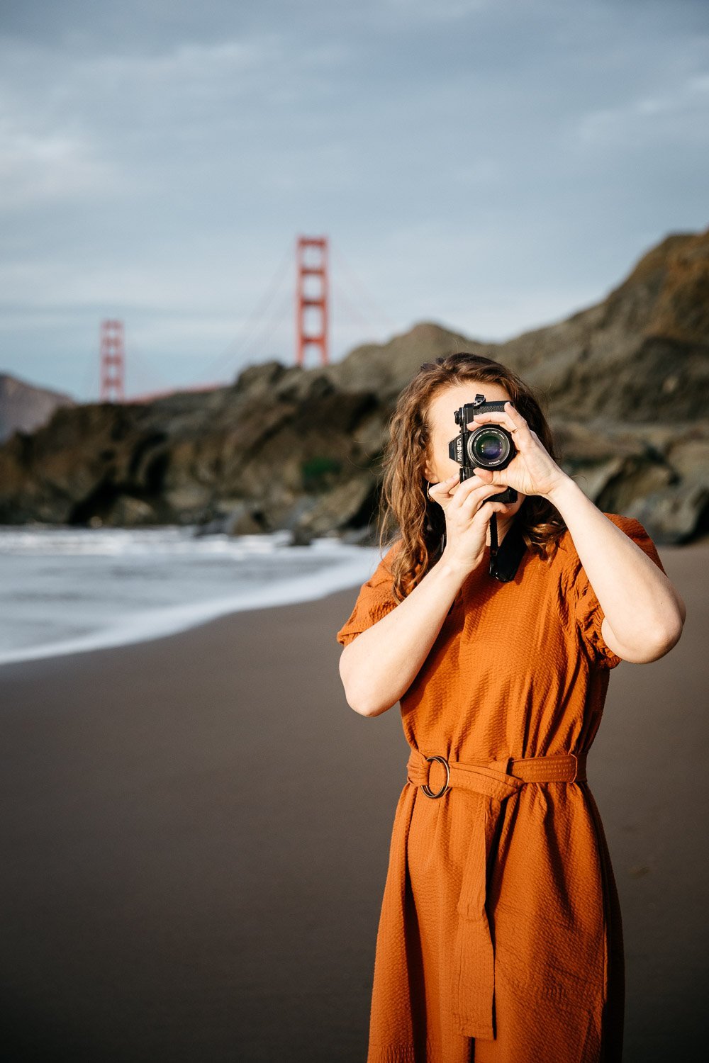  brand portrait of photographer Toni Toscano holding her Olympus camera with a cliff and the Golden Gate Bridge behind her, shot at Baker Beach in San Francisco by brand photographer Allison Busch Photography 