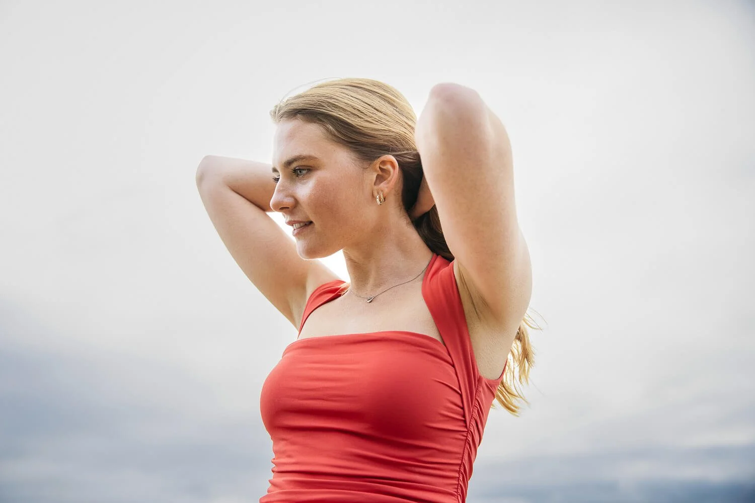 Teen girl wearing coral-colored tank top pulls back long hair back and looks off in the distance, shot during senior session at Ocean Beach.