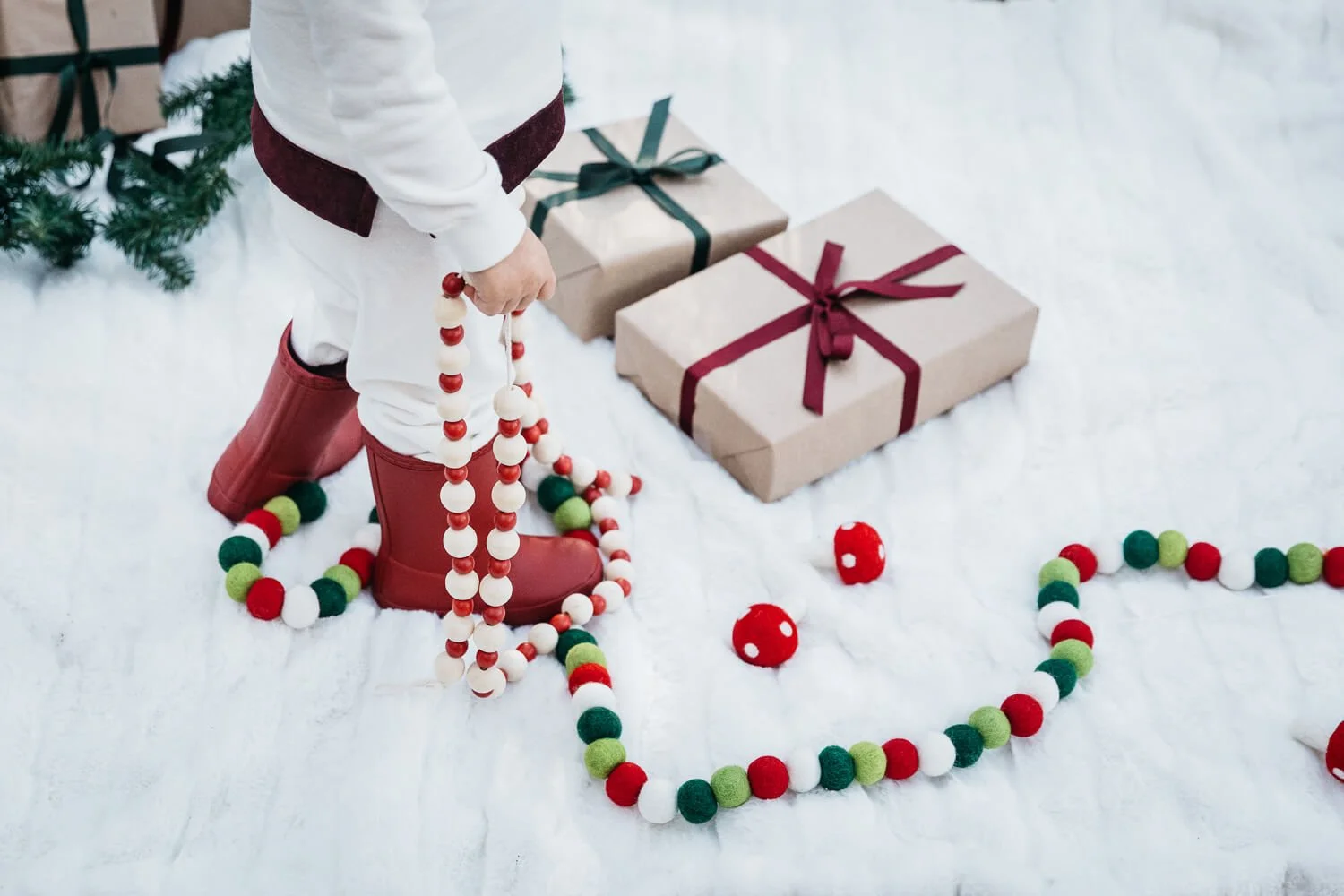 Crop of toddler holding christams garland while standing on white blanket, surrounded by presents.