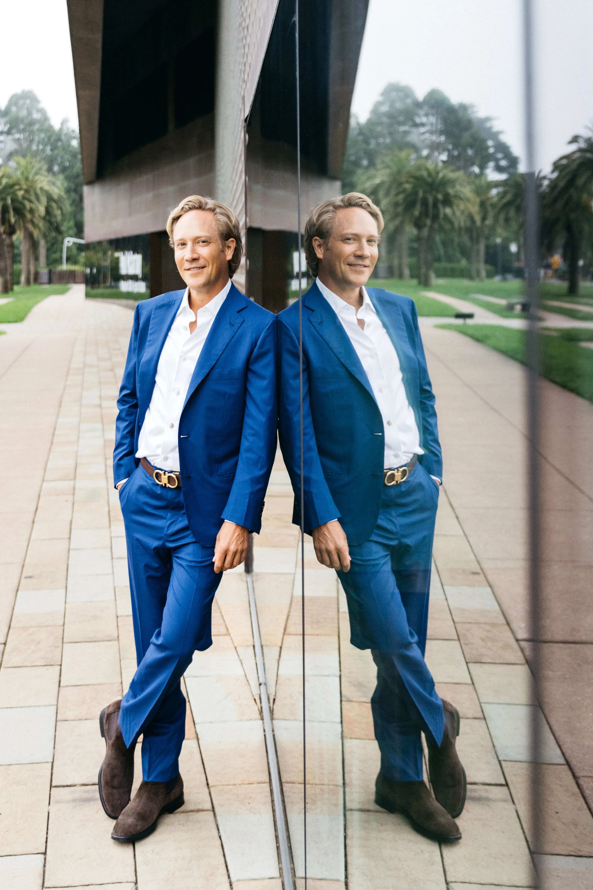 Man in blue suit with white shirt leans against reflective glass wall at the deYoung Museum in San Francisco.