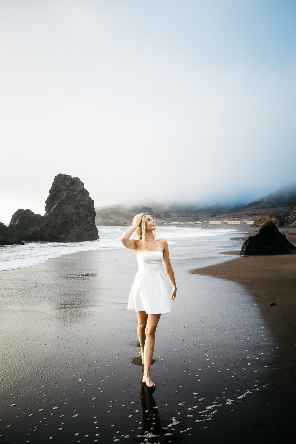 Teen girl with long hair and short white dress walks in surf at foggy Rodeo Beach, shot during senior session.